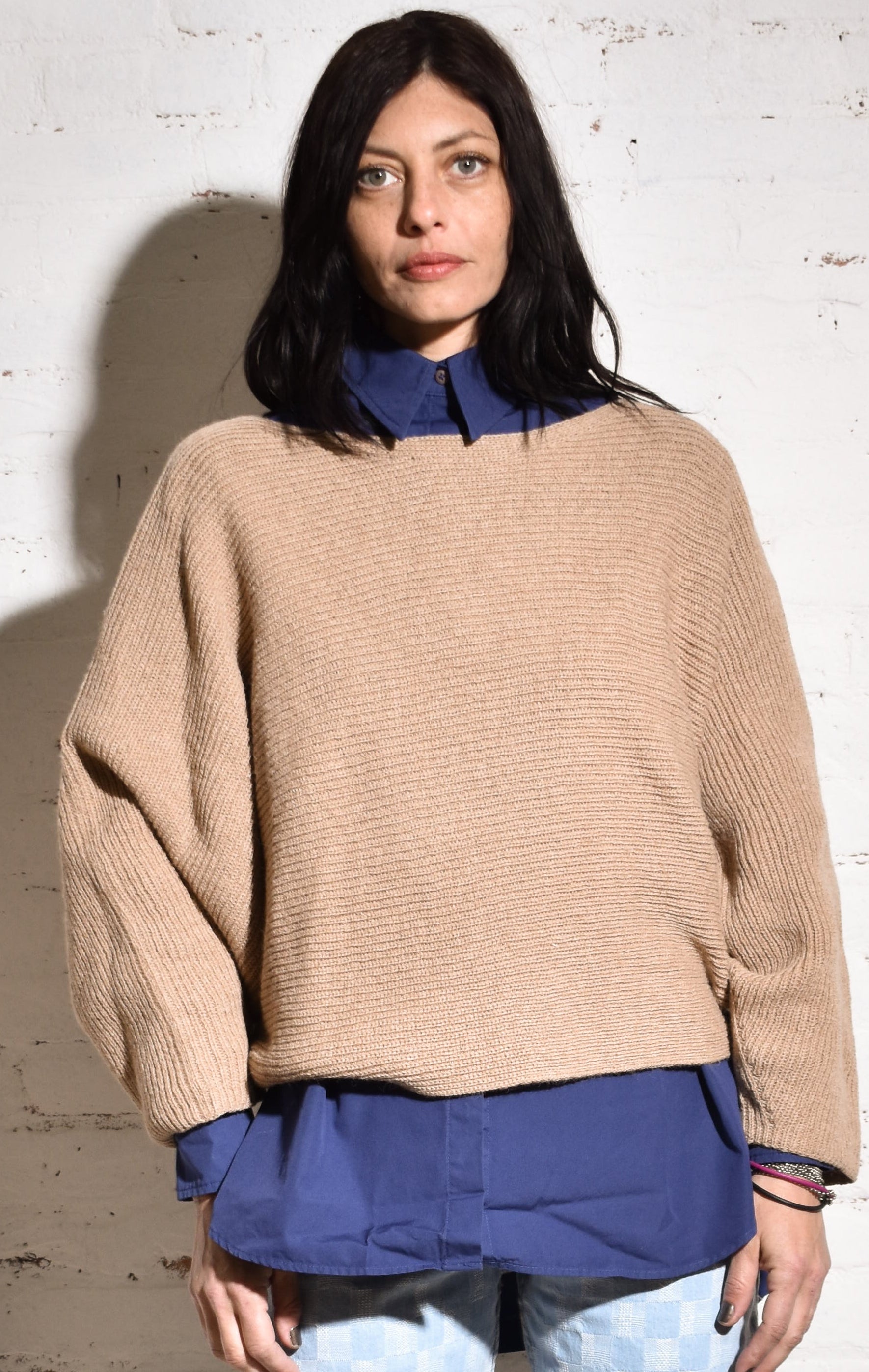 A person wearing a beige hand-knit alpaca sweater with a boat neck and batwing sleeves, paired with blue jeans and white sneakers.